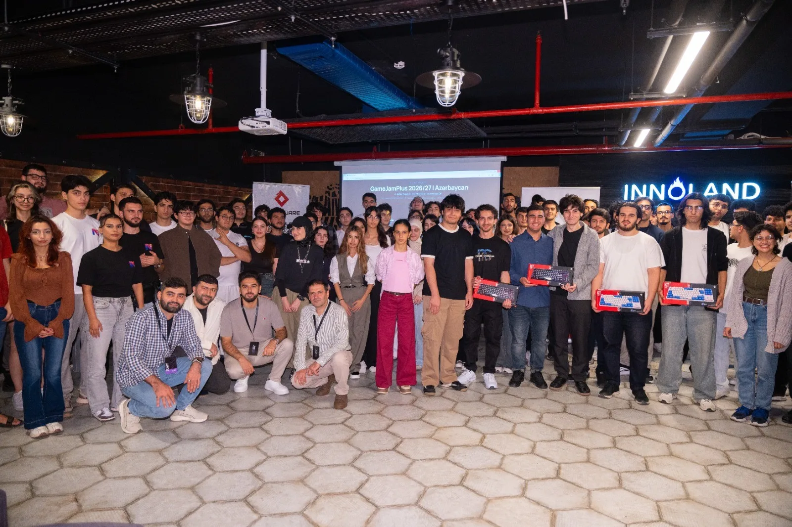 Group photo of GameJamPlus 2026/27 Azerbaijan participants, jury and organizers at INNOLAND, winners holding keyboard prizes, the event page on the projector behind them